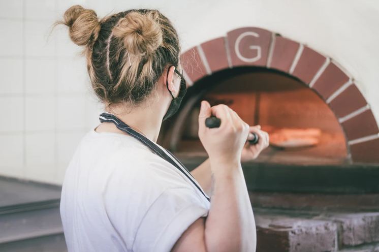 Image of a woman baking pizza in a wood-fired pizza oven.