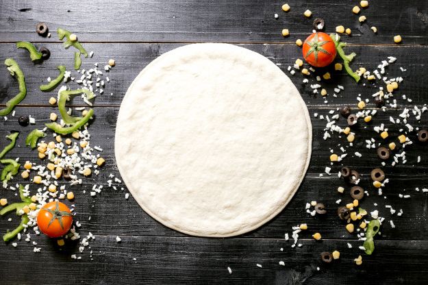 Picture of a pizza dough next to a variety of ingredients with a black wooden background.