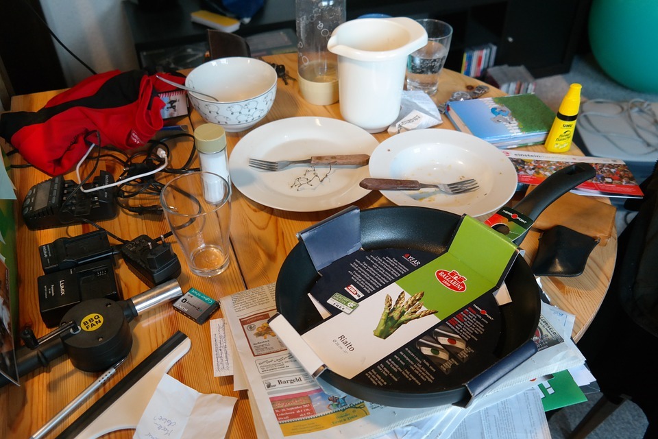 dining table, clutter, frying pan, glasses, plates, saucer, forks, battery, pedals, salt shaker, papers, piece of clothing