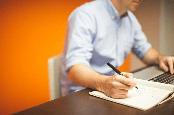 man working, pen, orange wall, open notebook, laptop, chair