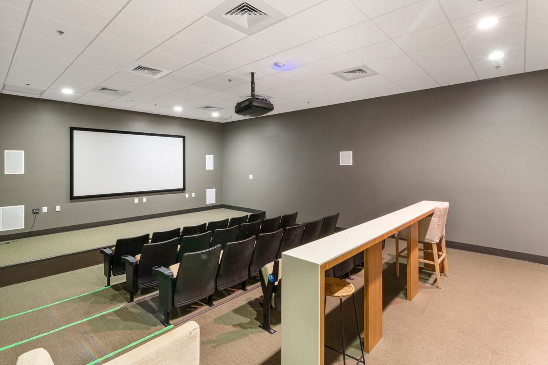 an empty classroom with a white scree, chairs, projector, table, and grid ceilings