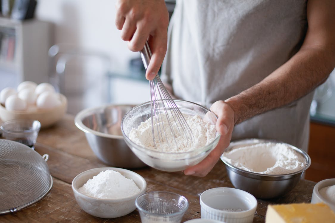 a man holding a bowl of flour and using a whisk to sift it, baking ingredients on the table