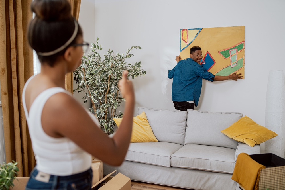 Couple in love decorating their first apartment, unpacking boxes after moving in, the man hang a picture on the wall, the woman shows her thumb up as a sign that it looks good and hangs evenly